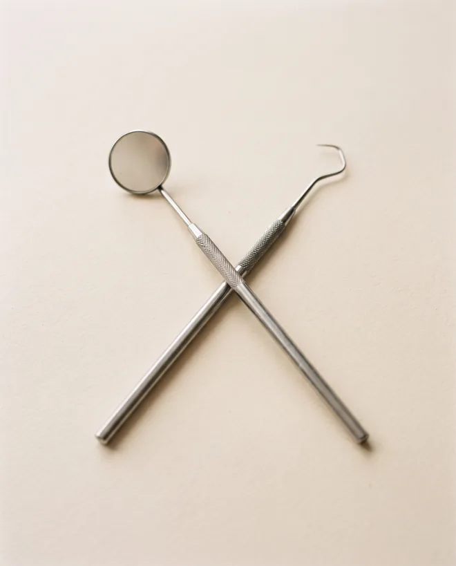 A dental mouth mirror and a sickle explorer laid crossed on warm off-white paper