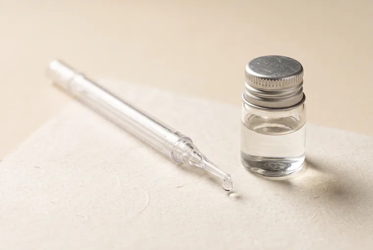 A slim clear plastic dental applicator with a fine tapered tip lying diagonally on warm off-white paper beside a small clear glass vial with a brushed silver cap