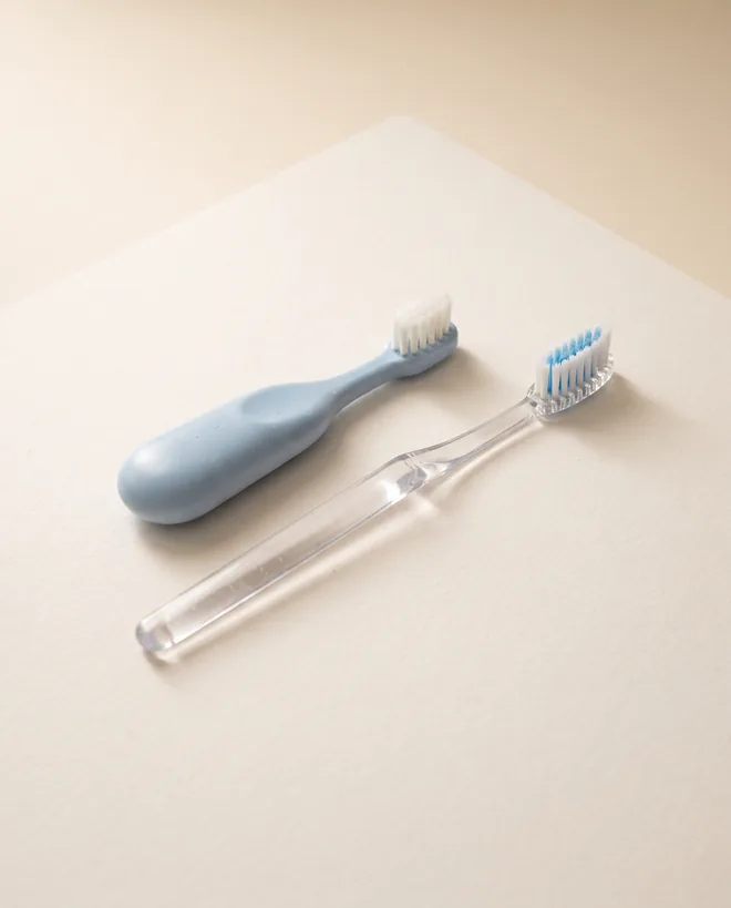 A small child-sized toothbrush with a short chunky handle lying next to a full-sized adult toothbrush on warm off-white paper