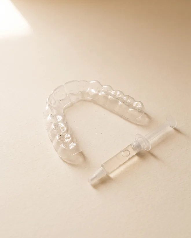 A clear custom-fitted whitening tray lying on warm off-white paper beside a small syringe of pale whitening gel